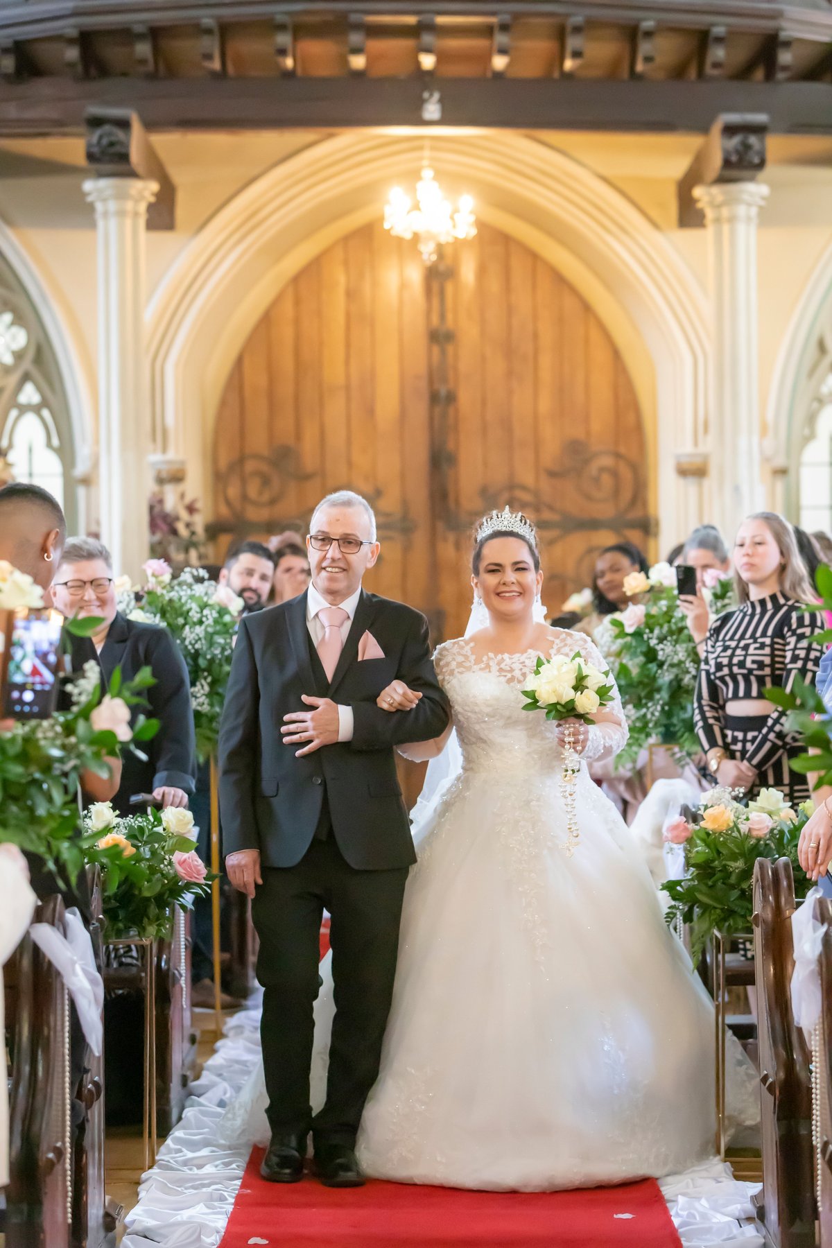Noiva e seu pai caminhando pelo corredor da igreja decorado com flores brancas e folhagens verdes