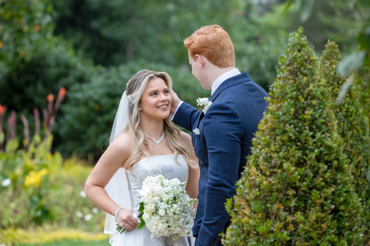 Noiva de vestido branco e véu segurando flores brancas olha para o noivo de terno azul em jardim