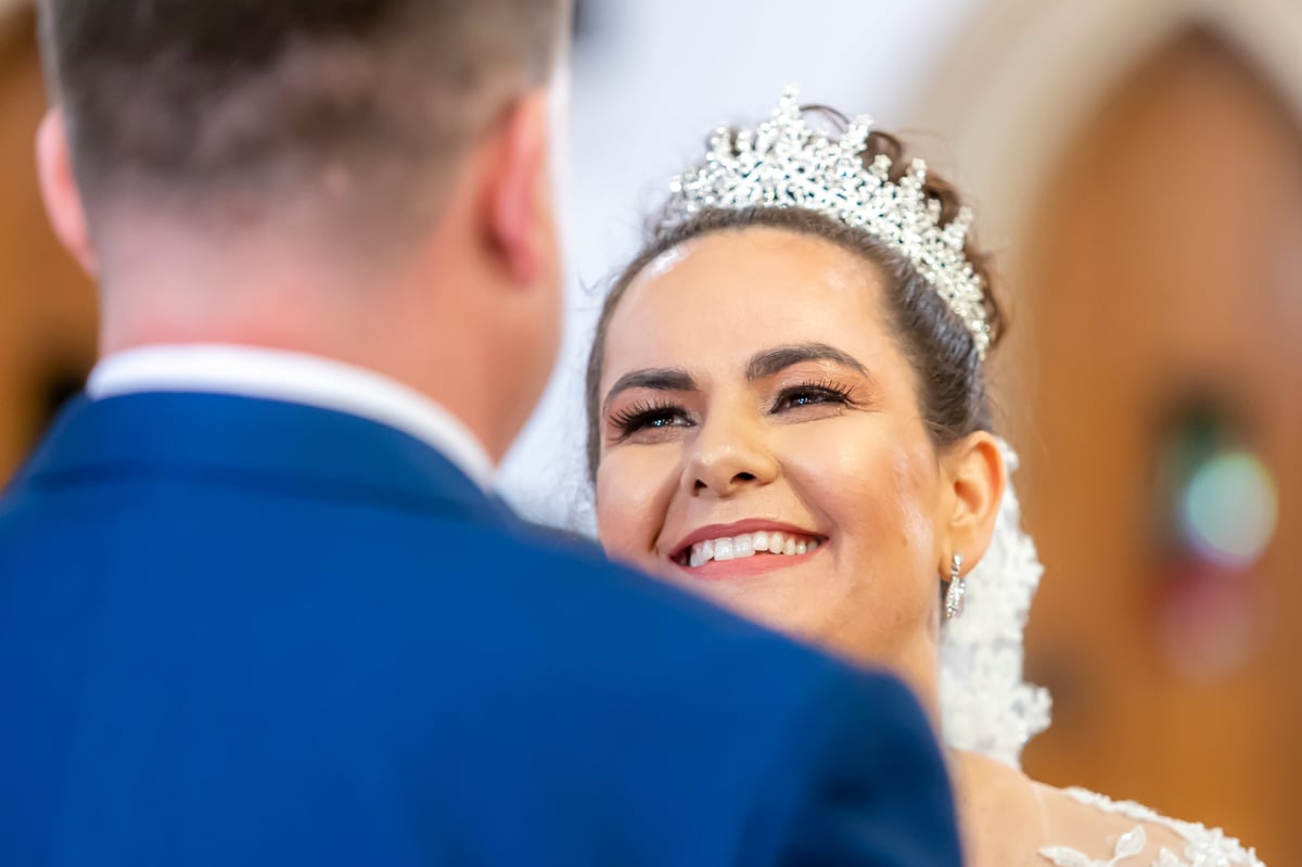 Noiva de véu branco rendado e tiara sorrindo para o noivo de terno azul durante a cerimônia