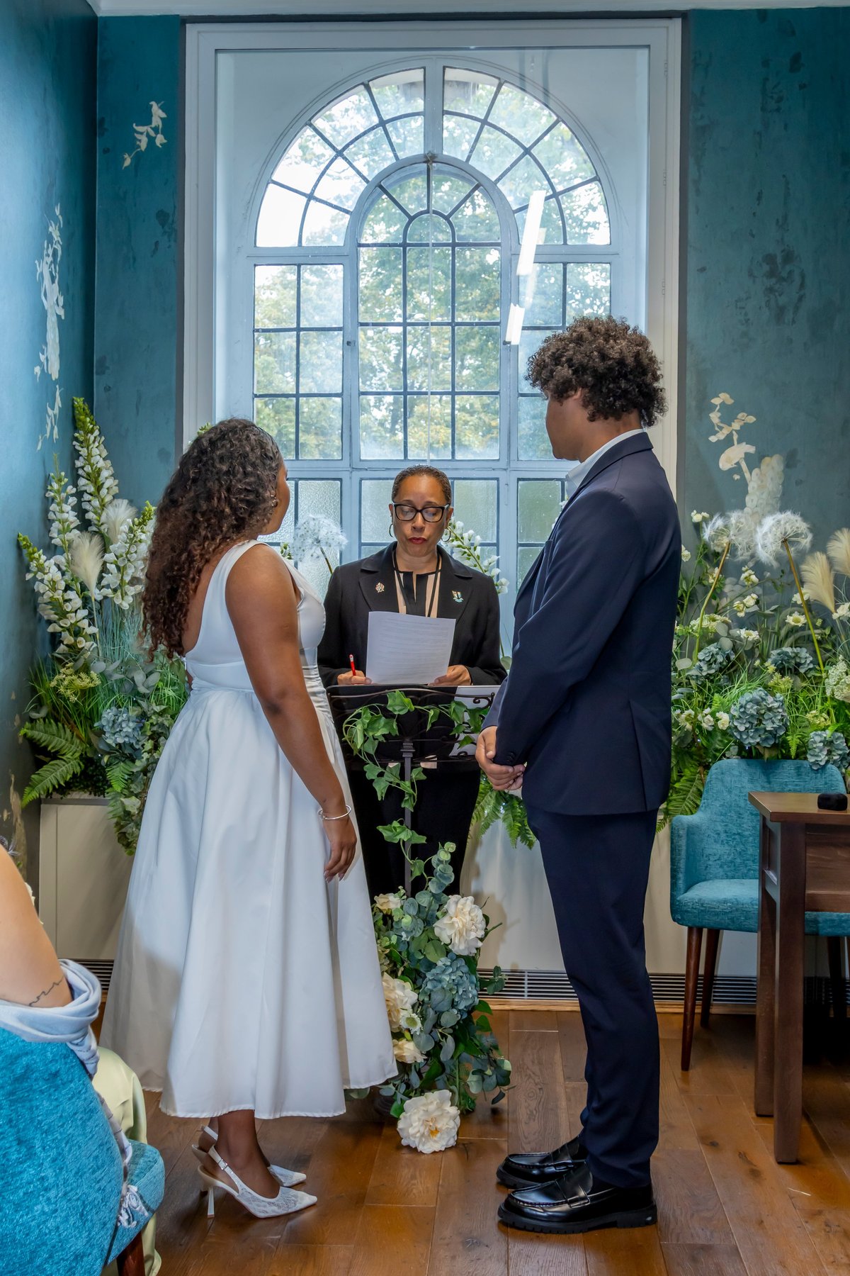 Cerimônia de casamento com noiva de vestido branco, noivo de terno azul-marinho e celebrante diante da janela em arco