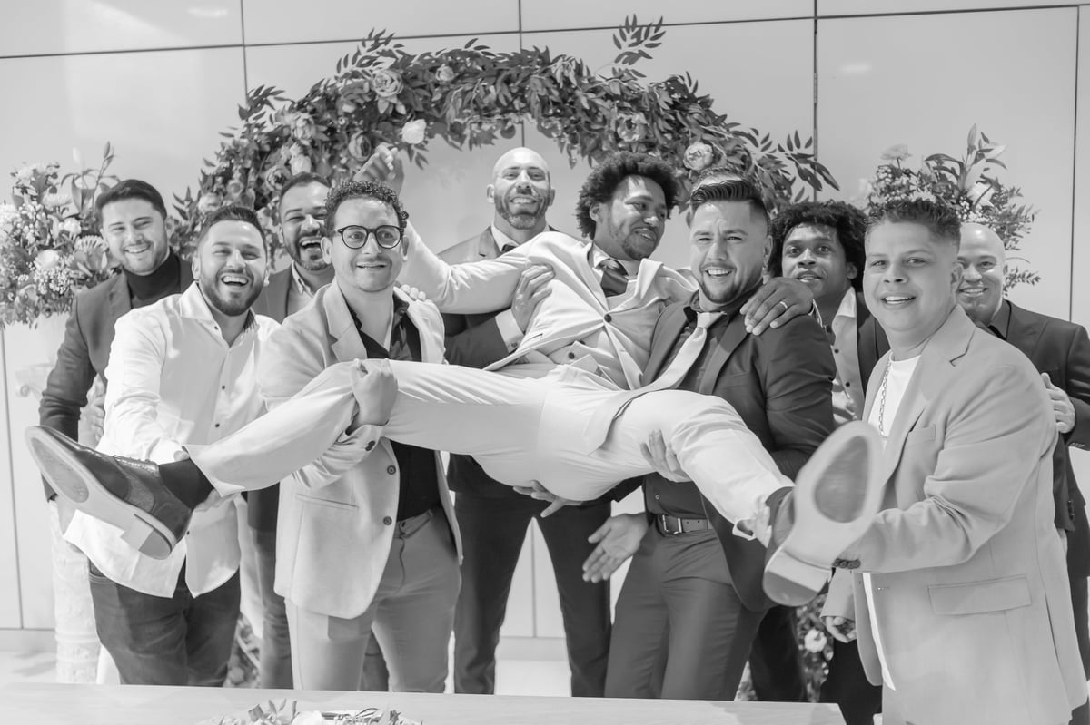 Foto em preto e branco de grupo diverso de homens de terno erguendo a noiva no colo, celebrando sob arco floral