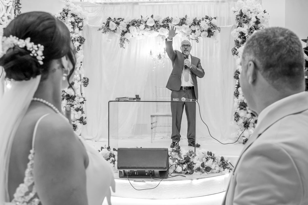 Foto em preto e branco dos noivos observando o celebrante sob o arco decorado com arranjos florais