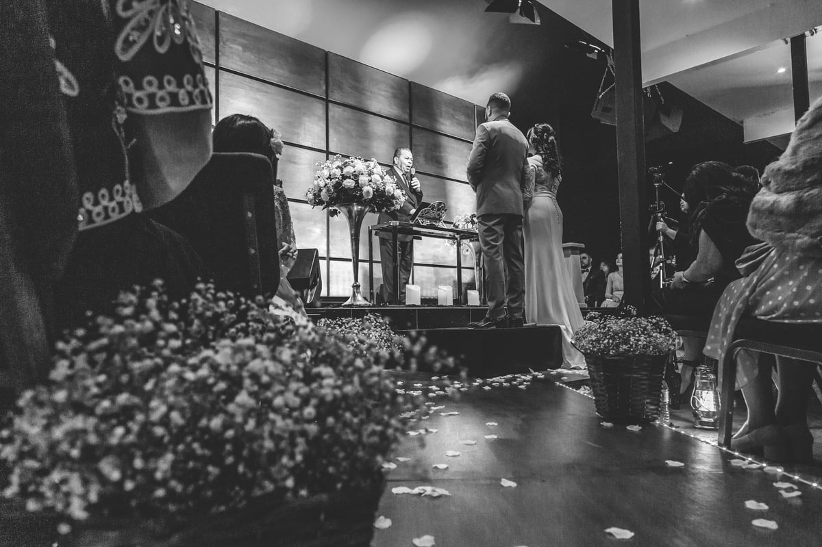 Foto em preto e branco da cerimônia de casamento com noiva e noivo no altar cercados por arranjos florais e convidados