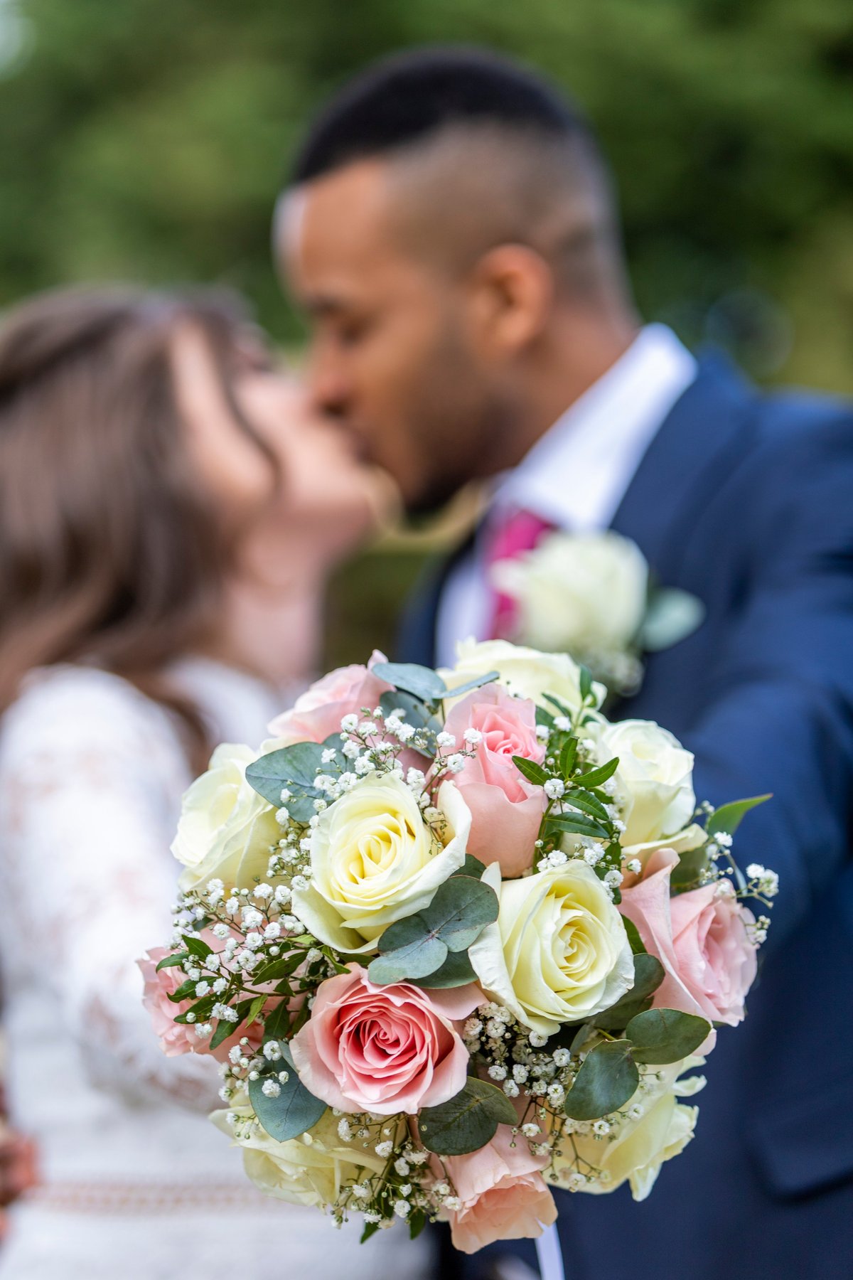 Buquê de casamento com rosas brancas, amarelas e rosadas e mosquitinho, com casal se beijando desfocado ao fundo